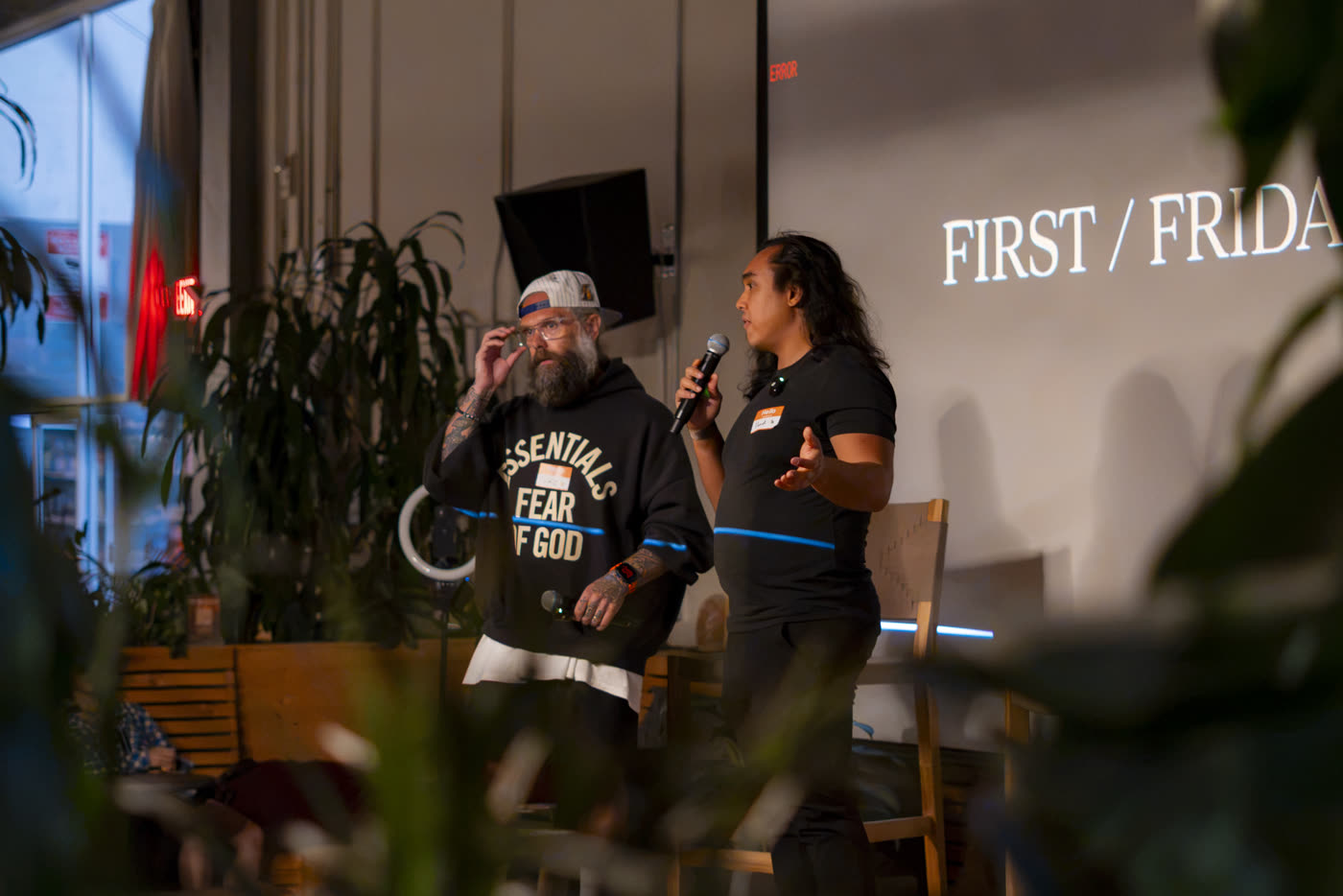 Two hosts on stage with a mic in front of the FIRST/FRIDAYS slide